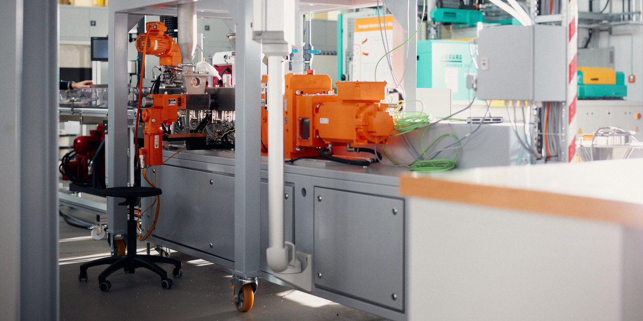 Bild des Doppelschneckenextruders mit seinen verschiedenen Komponenten Im Vordergrund sieht man einen orangenen Elektromotor darunter das graue Gehäuse in dem die Drehstäbe eingebaut sind